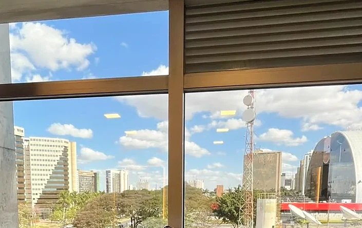 Vista da janela da sala de atendimento: céu azul com nuvens, prédios e árvores da Asa Norte, em Brasília